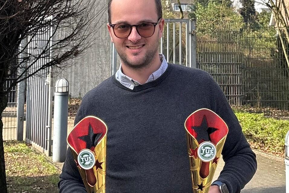 Mann mit Brille hält zwei goldene Pokale vor einem Zaun und Bäumen im Hintergrund.