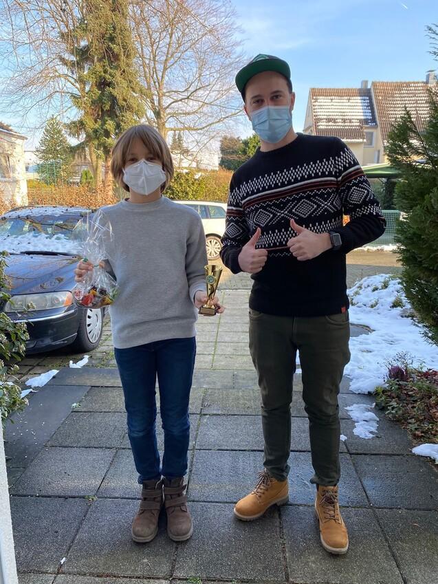 Zwei Personen stehen auf einem Gehweg, der mit Schnee bedeckt ist, und halten einen Pokal sowie eine Tüte mit Süßigkeiten.