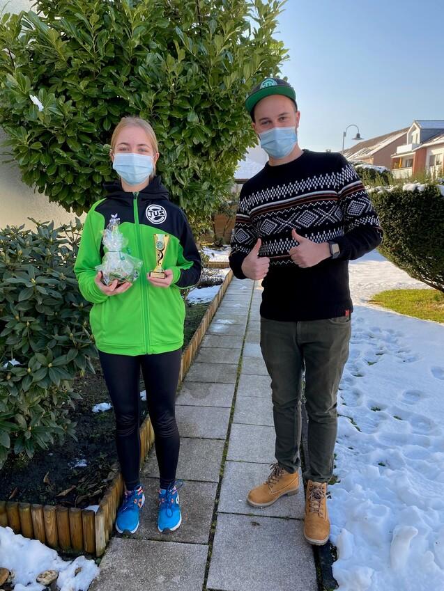 Zwei Personen stehen im Freien neben einer Hecke, eine hält einen Pokal und eine Tüte, beide tragen Masken.