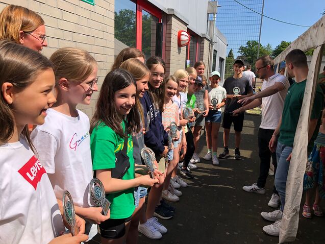 Gruppe von M&auml;dchen mit Pokalen stehen in einer Reihe, w&auml;hrend Erwachsene ihnen applaudieren und gratulieren.