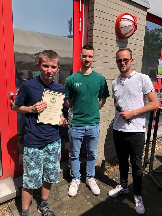 Drei junge M&auml;nner stehen vor einem Geb&auml;ude, einer h&auml;lt eine Urkunde und einen Pokal in der Hand.