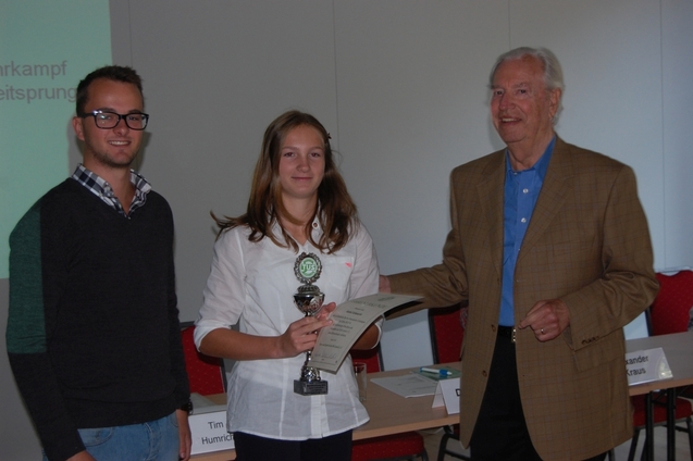 Drei Personen stehen an einem Tisch mit Urkunde und Pokal in einem Raum mit roten St&uuml;hlen im Hintergrund.