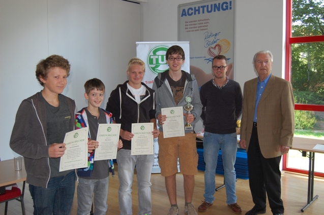 Gruppenszene mit vier Jugendlichen, die Urkunden halten, und einem &auml;lteren Mann neben einem Pokal in einem Raum.