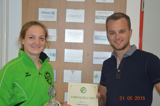 Zwei Personen, ein Mann und eine Frau, halten eine Ehrenurkunde vor einer Wand mit Sponsorenlogos.