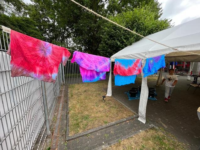 Bunte T-Shirts in lila und blau h&auml;ngen auf einer Leine vor einem Zelt in einem Garten.