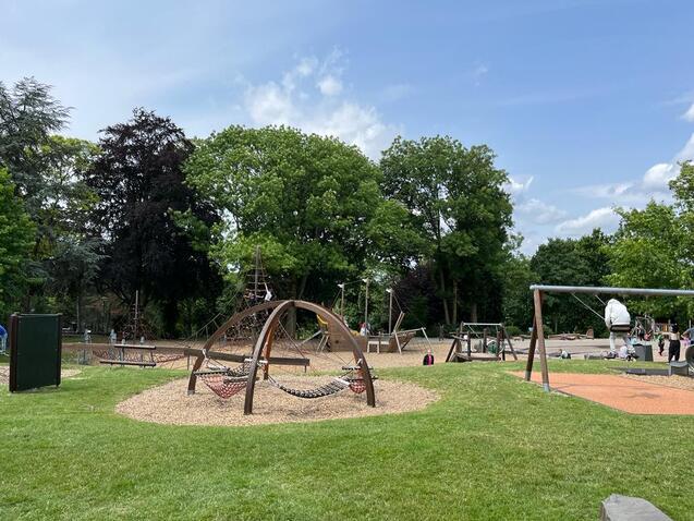 Spielplatz mit einer H&auml;ngematte und Schaukeln, umgeben von B&auml;umen und Rasenfl&auml;chen unter blauem Himmel.