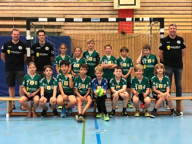 Gruppenspieler und Trainer einer Handballmannschaft in grünen Trikots posieren lächelnd vor einem Handballtor in einer Sporthalle.