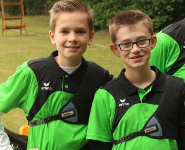 Zwei Jungen in gr&uuml;nen Polo-Shirts l&auml;cheln, tragen Sportausr&uuml;stung und stehen auf einer Rasenfl&auml;che im Freien.