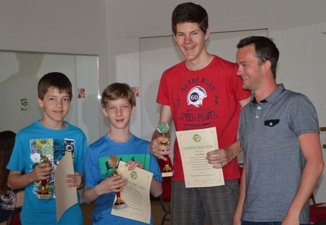 Drei Jugendliche halten Pokale und Urkunden, w&auml;hrend ein Erwachsener ihnen l&auml;chelnd gratuliert.