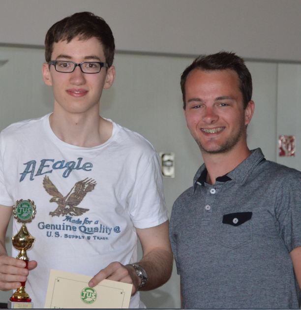 Zwei junge M&auml;nner posieren in einem Innenraum, einer h&auml;lt einen Pokal und ein Dokument in der Hand.