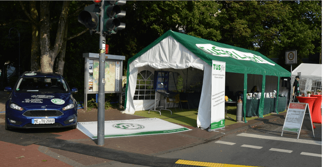 Blick auf einen Informationsstand mit grünem Zelt, Auto und Verkehrsampel an einer Straße.