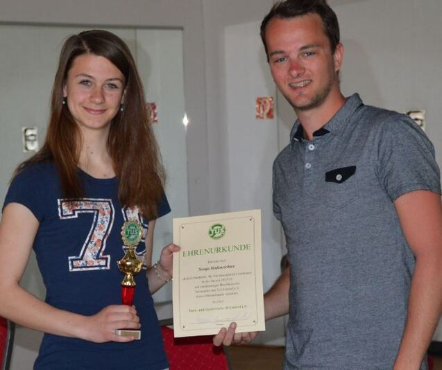 Eine junge Frau h&auml;lt einen Pokal und eine Urkunde, w&auml;hrend ein junger Mann daneben steht und l&auml;chelt.