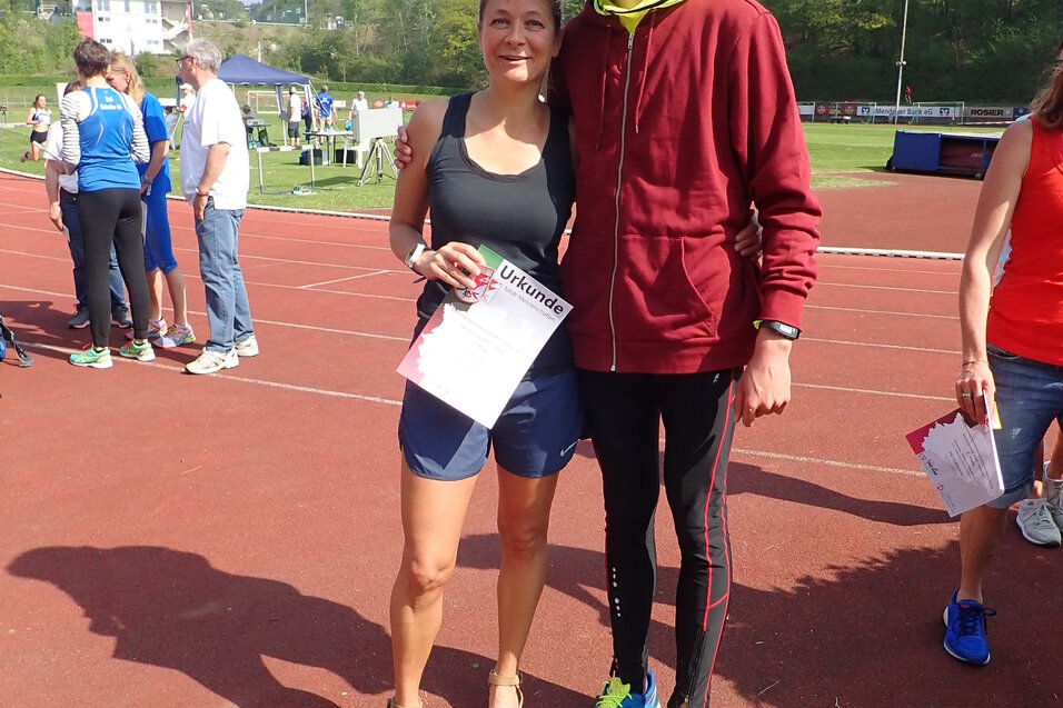 Zwei Personen posieren auf einer Laufbahn, eine h&auml;lt eine Urkunde, im Hintergrund sind weitere Teilnehmer sichtbar.