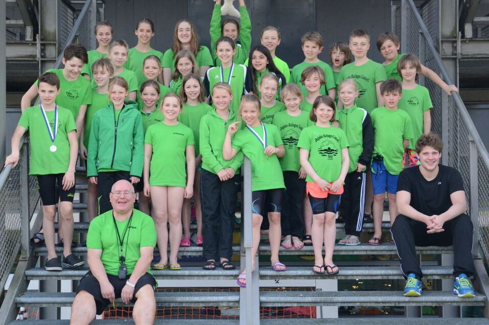 Gruppe von schwimmenden Kindern in gr&uuml;nen T-Shirts steht auf Treppen, einige halten Medaillen; ein Trainer sitzt vorne.
