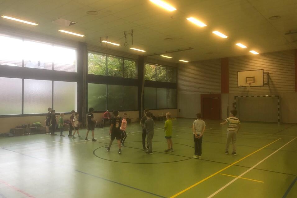 Gruppe von Kindern spielt in einer Sporthalle mit Basketballkorb, helle Fenster und gr&uuml;ne Bodenfl&auml;che.