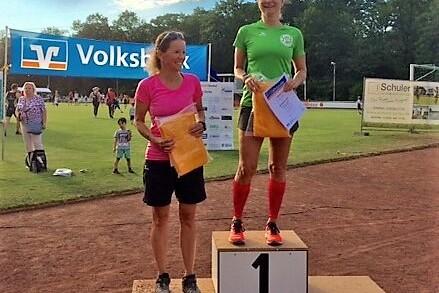 Zwei Frauen stehen auf dem Siegertreppchen: eine in Rot als Erste, eine in Pink als Zweite, mit Urkunden in der Hand.