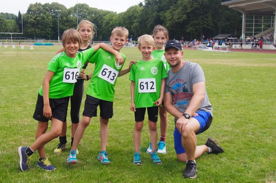 Gruppenbild von f&uuml;nf Kindern in gr&uuml;nen Sporttrikots mit Wettkampfnr. 612, 613, 616, zusammen mit einem Trainer auf einer Wiese.