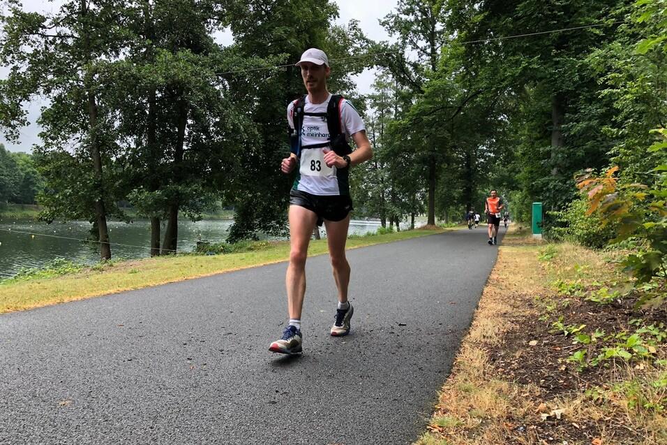 L&auml;ufer in sportlicher Kleidung mit Nummer 83 joggt auf einem Weg am See, umgeben von B&auml;umen und Natur.