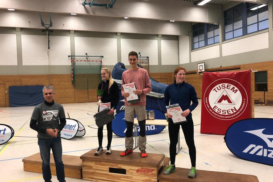 Drei Sportler stehen auf dem Siegertreppchen und halten Urkunden in einer Sporthalle, mit Sponsorenbannern im Hintergrund.