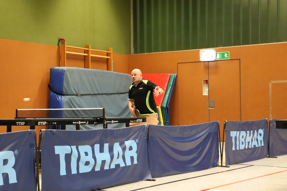 Aktiver Tischtennisspieler schl&auml;gt den Ball &uuml;ber das Netz in einer Sporthalle mit Tischtennisplatten und Matten im Hintergrund.