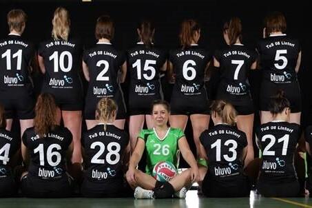 Gruppenfoto einer Handballmannschaft mit Spielerinnen in schwarzen Trikots, die R&uuml;ckennummern zeigen, eine Spielerin in gr&uuml;n sitzt vorne.