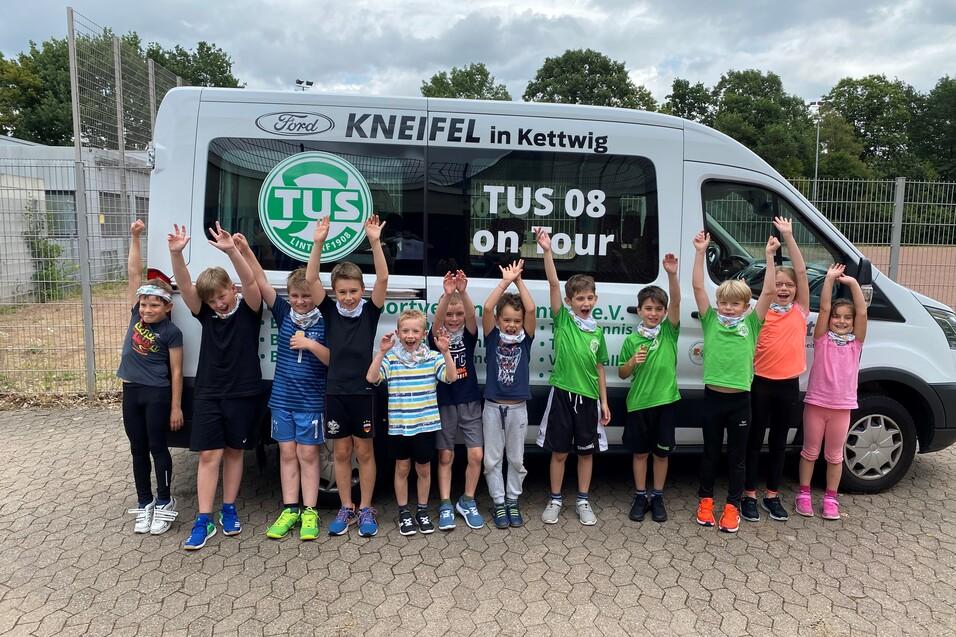 Gruppe von Kindern in Sportkleidung steht vor einem Vereinsbus mit "TUS 08 on tour" Aufdruck und hebt die H&auml;nde.