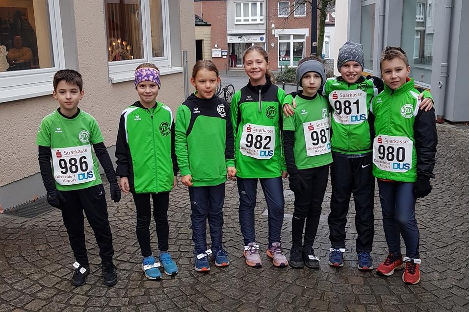 Gruppe von sieben Kindern in gr&uuml;nen Sportshirts und Startnummern auf einem Kopfsteinpflasterweg vor Geb&auml;uden.