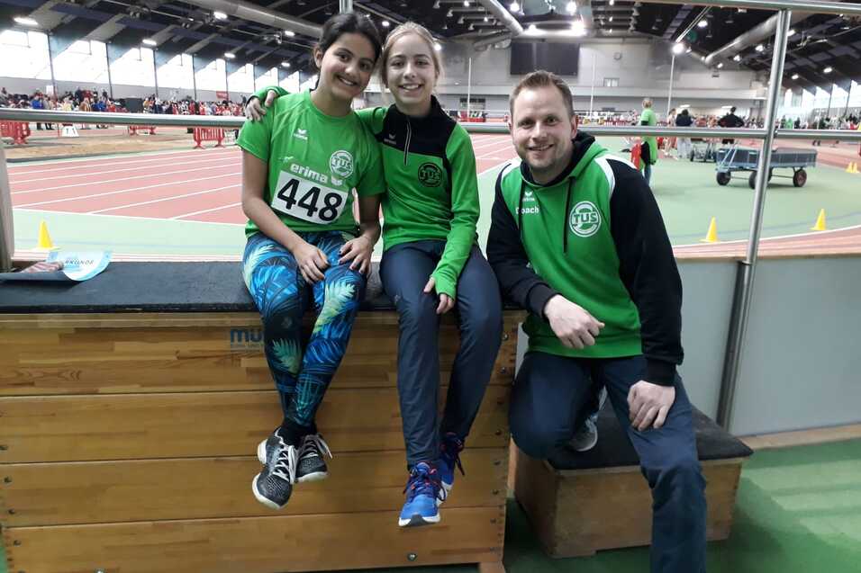 Drei Personen, zwei M&auml;dchen und ein Junge, in Sportoutfits sitzen auf einer Holzbank in einer Sporthalle.
