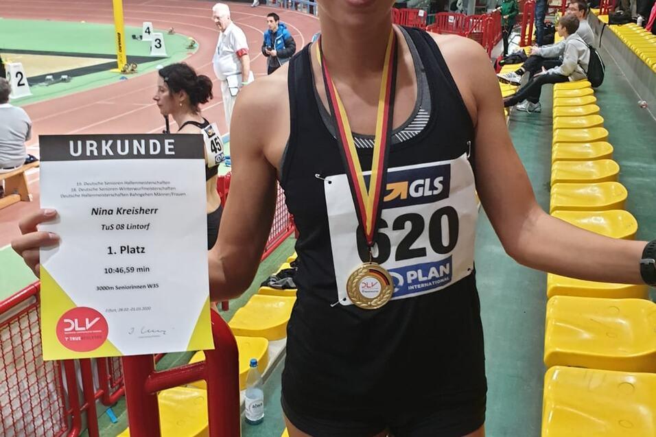 Sportlerin mit Medaille und Urkunden in der Hand, steht in einer Sporthalle mit gelben Sitzen im Hintergrund.