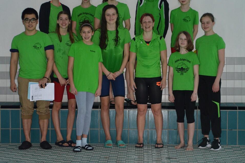 Gruppe von Schwimmern in gr&uuml;nen Shirts steht am Beckenrand eines Schwimmbades. Im Vordergrund Wasseroberfl&auml;che sichtbar.