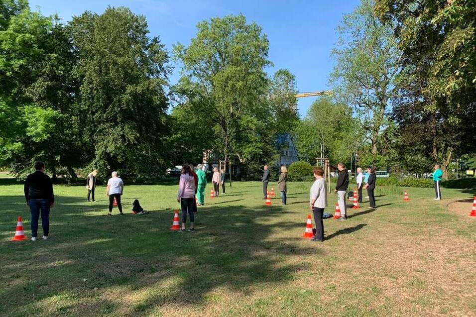 Gruppe von Menschen in einem Park, die Abstand halten, um orangefarbene Pylonen herum auf einer Wiese stehen.
