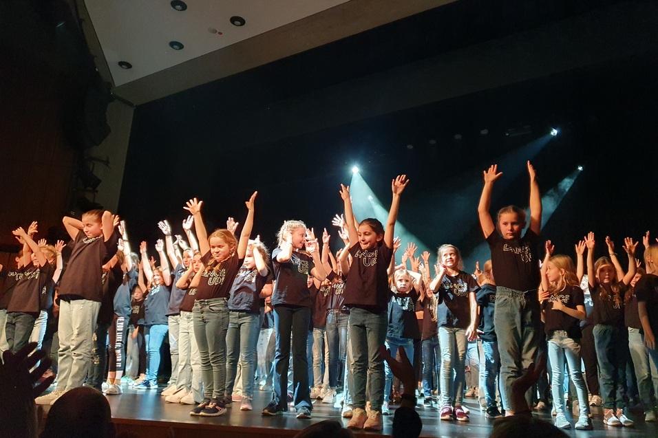 Gruppendarbiet von Kindern auf einer B&uuml;hne, die fr&ouml;hlich mit erhobenen H&auml;nden tanzen.