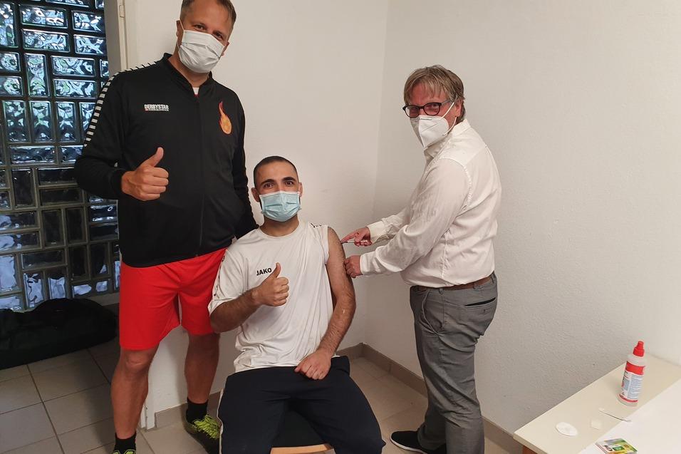 Zwei M&auml;nner mit Mund-Nasen-Schutz zeigen Daumen hoch, w&auml;hrend ein Dritter impft. Der Geimpfte sitzt auf einem Stuhl.