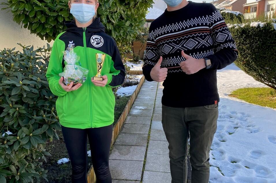 Zwei Personen stehen im Freien neben einer Hecke, eine h&auml;lt einen Pokal und eine T&uuml;te, beide tragen Masken.