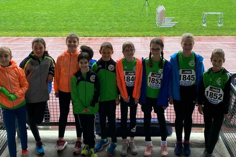 Gruppe von neun jungen Athleten in Sportkleidung mit Startnummern, stehend auf der Trib&uuml;ne eines Sportplatzes.