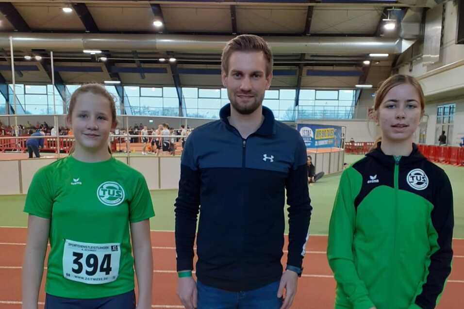 Drei Sportlerinnen stehen in einem Hallenrennen, zwei in gr&uuml;nen Trikots, ein Trainer zwischen ihnen.