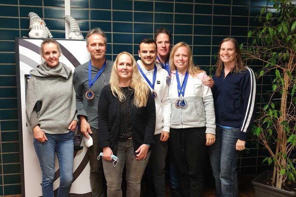 Sechs Personen mit Medaillen stehen vor einem Getr&auml;nkeautomaten in einem Flur mit gr&uuml;nem Fliesenmuster.