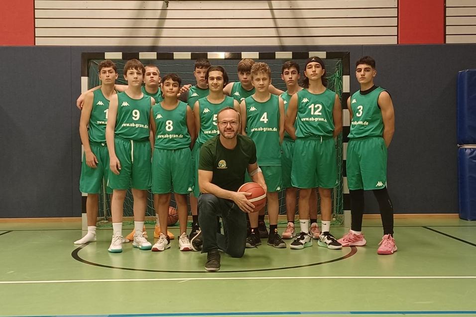 Basketballmannschaft in gr&uuml;nen Trikots posiert gemeinsam auf dem Spielfeld, ein Trainer h&auml;lt den Ball in der Mitte.