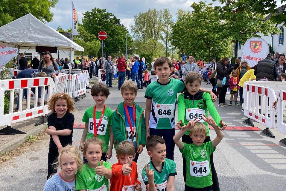 Gruppeneinstellung von sieben Kindern in Sportkleidung mit Medaillen nach einem Laufwettbewerb, im Hintergrund eine Startbanner.