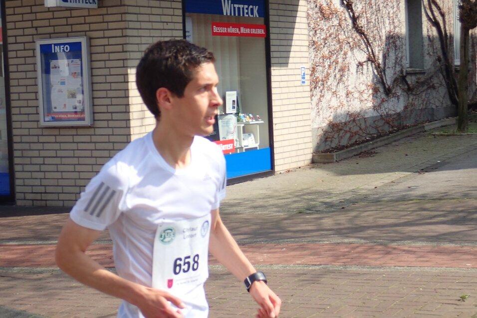 L&auml;ufer in wei&szlig;em Shirt mit Startnummer 658 joggt an einem Gesch&auml;ft vorbei. Helle, sonnige Stra&szlig;enumgebung.