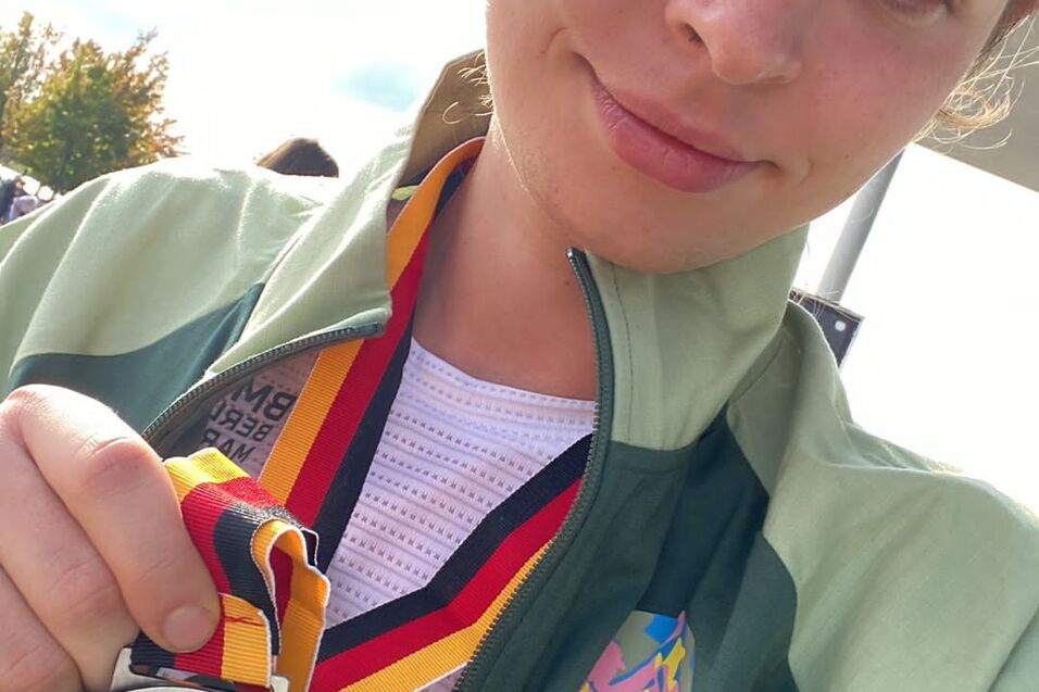 Junge Athletin h&auml;lt eine silberne Medaille mit rot-gelb-schwarzem Band vor ihrem Gesicht, im Sportoutfit mit Jacke.