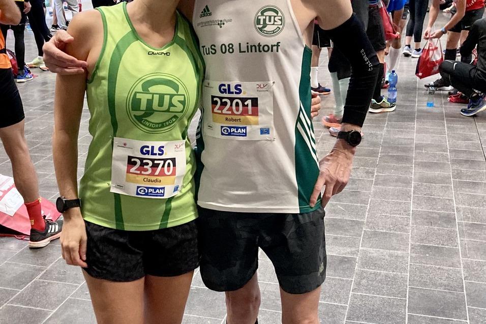 Zwei L&auml;ufer stehen in Sportbekleidung mit Startnummern in einer Halle voller Teilnehmer und Vorbereitungen.