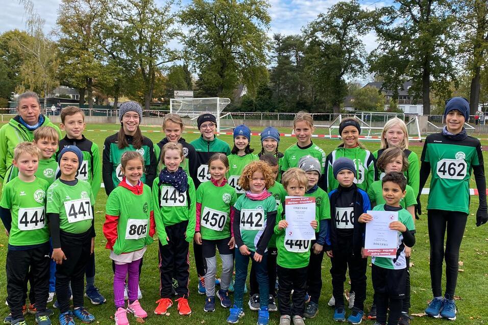 Gruppe von 25 Kindern in gr&uuml;nen Trikots mit Startnummern auf einem Sportplatz, einige halten Urkunden in den H&auml;nden.