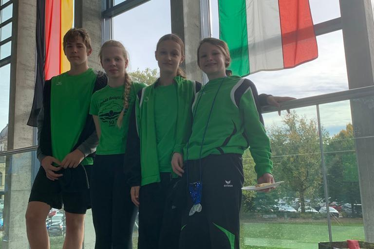 Vier Kinder in gr&uuml;nen Sportanz&uuml;gen stehen an einem Fenster mit Flaggen im Hintergrund.