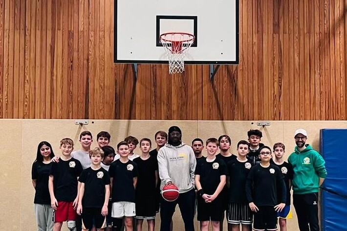 Gruppe von Jugendlichen in Basketballtrikots steht vor einem Basketballkorb in einer Sporthalle.