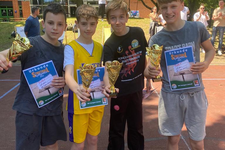 Vier Jungen halten goldene Pokale und Urkunden in der Hand, l&auml;cheln, vor einem Veranstaltungszelt auf einem Sportplatz.