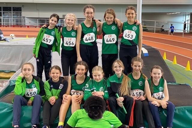 Gruppenzusammenstellung von zw&ouml;lf jungen Athletinnen in Sportkleidung mit Nummern auf einer Leichtathletikanlage.