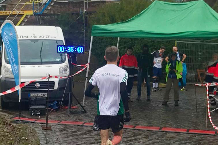 L&auml;ufer mit Nummer hinterlegt, sprintet auf einer gepflasterten Stra&szlig;e zur Zielmarkierung vor einer Versorgungsstation.