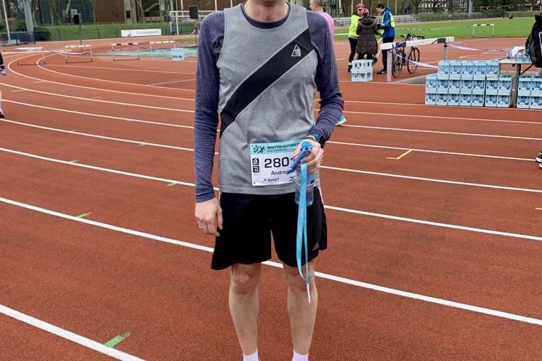 Mann in Sportkleidung mit Medaille steht auf einer Laufbahn, im Hintergrund sind andere L&auml;ufer und Geb&auml;ude sichtbar.