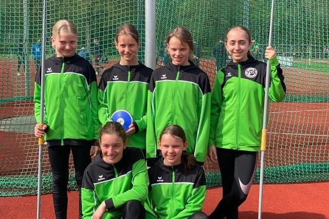 Sechs M&auml;dchen in gr&uuml;nen Sportjacken stehen und sitzen vor einer Sportanlage mit Wurfger&auml;ten.